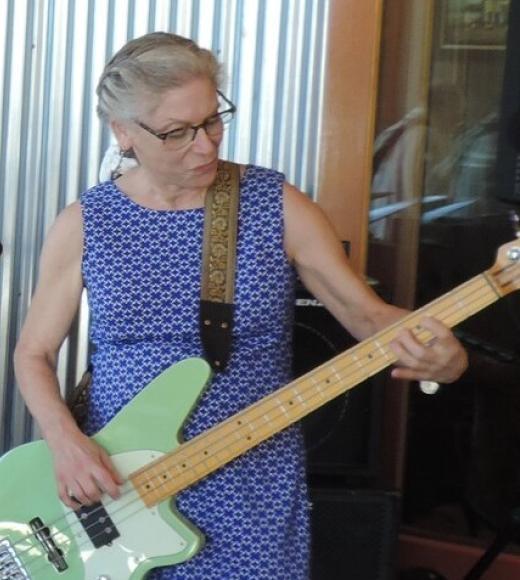 A woman in a blue dress and glasses plays an electric bass guitar.